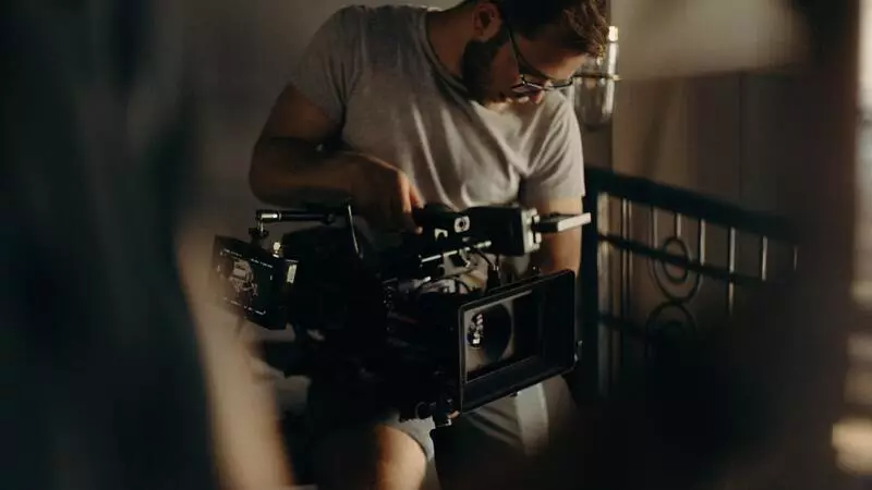 A videographer in a white t-shirt and glasses holding a large video camera on a video production shoot. 